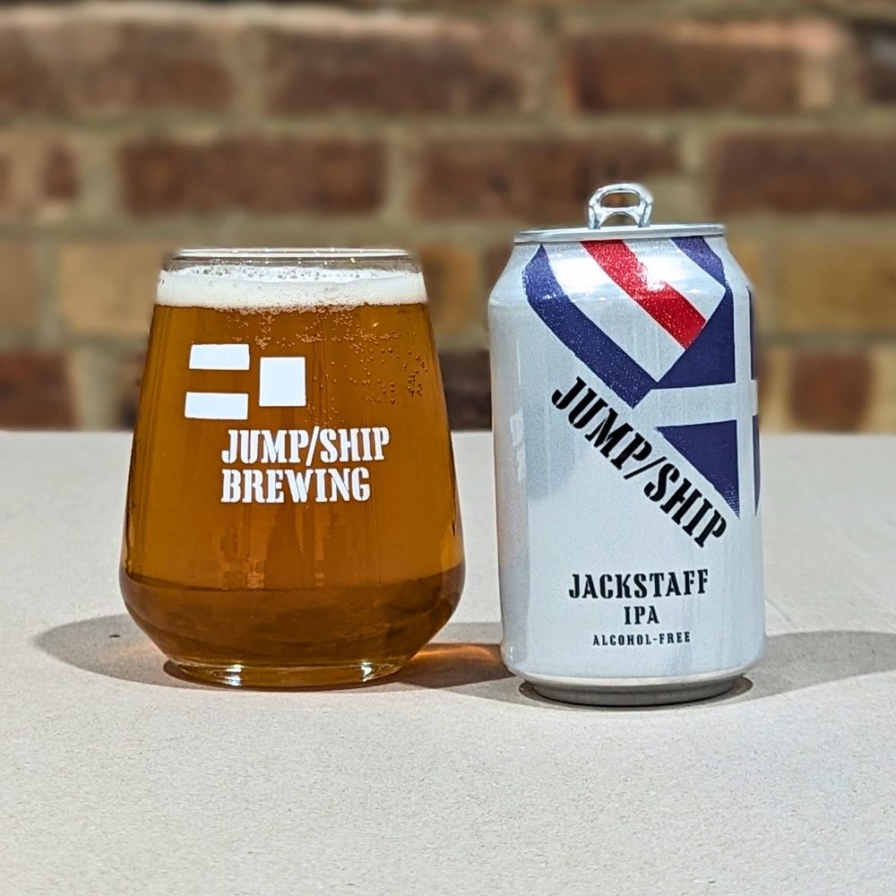 Glass and can of Jackstaff IPA from Jump/Ship Brewing on a table with a brick wall background.