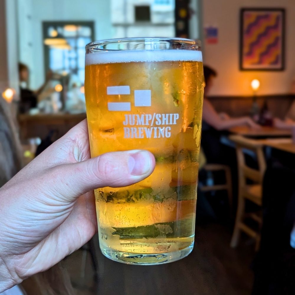 Glass of beer from Jump/Ship Brewing held in a hand with a blurred bar background
