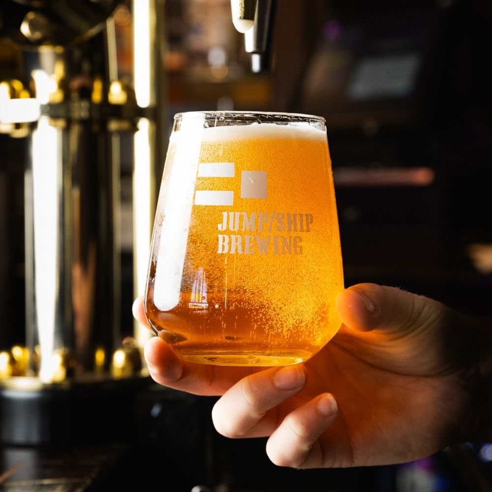 Hand holding a glass of beer with 'Jump ship Brewing' logo, blurred bar background