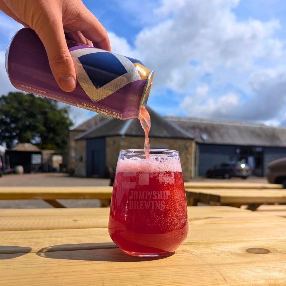 Person pouring a can of Jump Ship Brewing Ocean Drift Bramble Sour beer into a glass on a wooden table outdoors.
