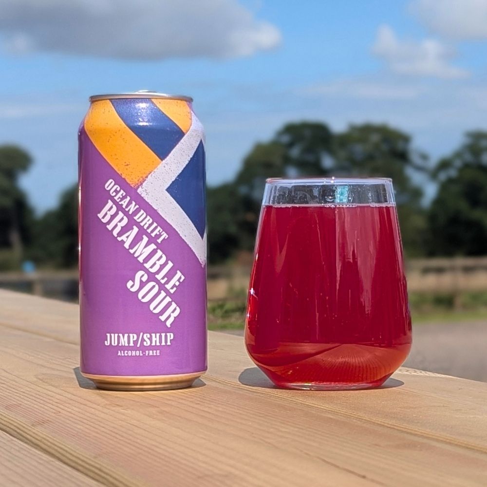 Can of Ocean Drift Bramble Sour next to a glass on a wooden surface with a blurred outdoor background.