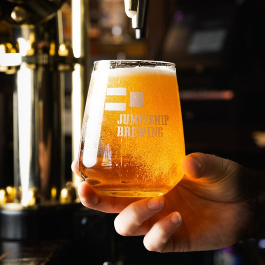 Hand holding a glass of beer with 'Jump ship Brewing' logo, blurred bar background